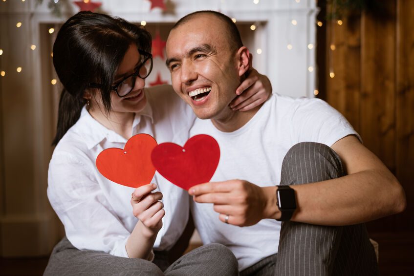 A couple smiling and laughing holding two paper hearts while sitting together